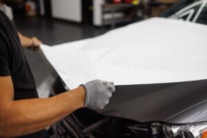 Person applying vinyl wrap to car hood in garage setting.